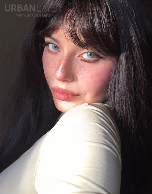 Close-up of a person with dark hair and freckles, light blue green contacts lenses and wearing a light-colored top.