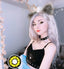 A person with long silver hair, cat ears, and a black gothic-style top poses indoors while wearing a bright yellow and black contact lens. A circular inset shows the lens design clearly.