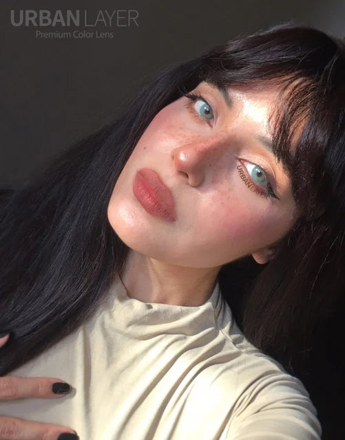 A close-up selfie of a woman with long dark hair, freckles, and light blue eyes wearing a cream-colored turtleneck. The image has a warm, stylized color cast and features the text 'URBAN LAYER Premium Color Lens' in the top-left corner.