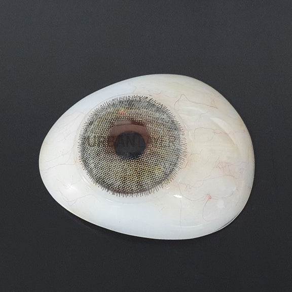 A realistic white eye contact lens prosthetic with a detailed gray and black iris, shown on a dark gray background.